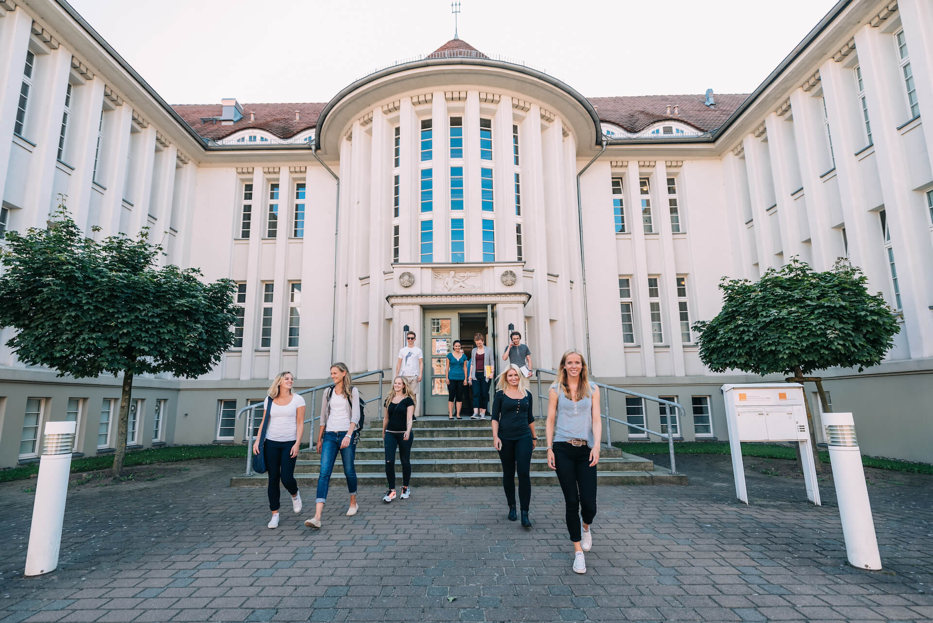 Wir sehen frontal auf den Standort Rostock. Junge, zufriedene Menschen verlassen das Gebäude durch das Hauptportal.